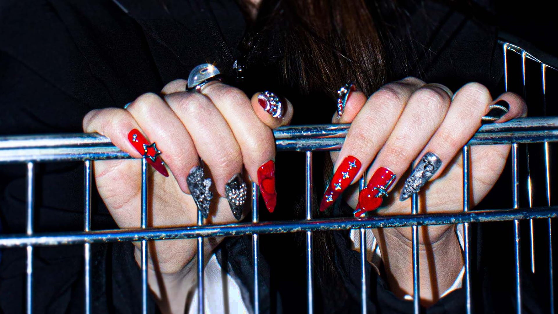 Close-up of hands with red press-on nails, polish and silver rings holding onto a metal bar.