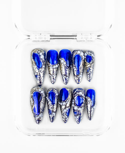 This press-on nails set features a striking, high-contrast aesthetic on sharp stiletto-shaped nails. The design is characterized by vibrant royal blue bases encased in heavily textured 3D silver "molten metal" frames that mimic flowing mercury. Scattered pearls of varying sizes are embedded into the silver textures, adding a touch of oceanic luxury to the edgy, sculptural hardware.