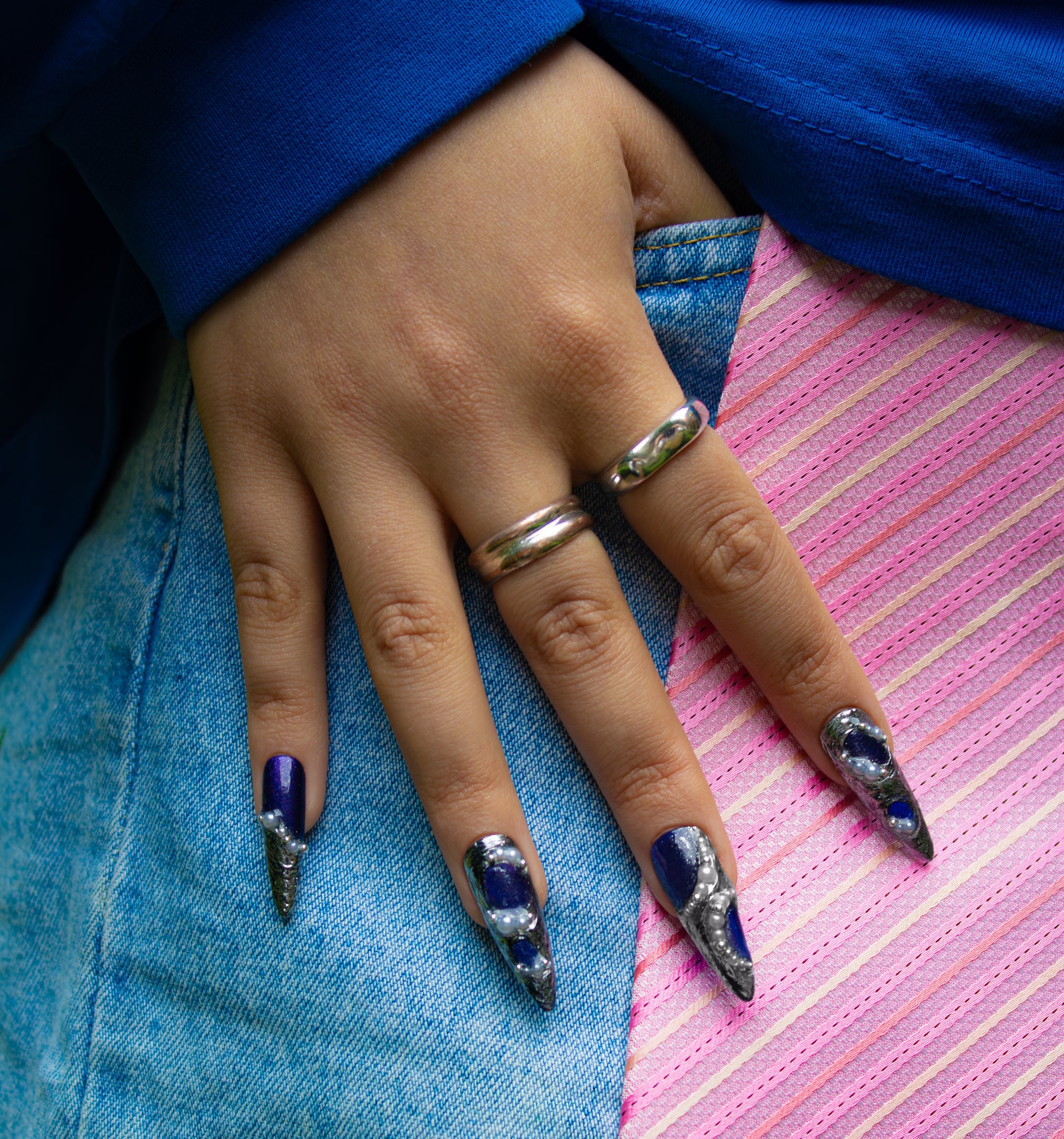 Hand with long, stiletto nails, the midnight blue set, with rings on a pink striped tie background