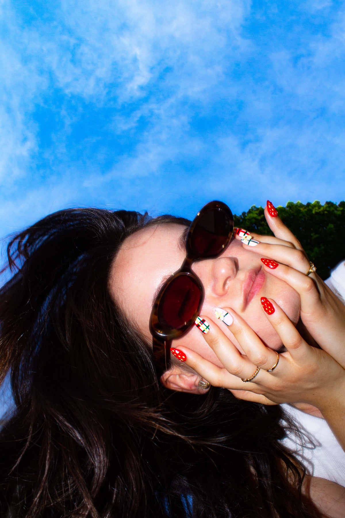 Model lying in grass in sunglasses, framing her face with strawberry press-on nails