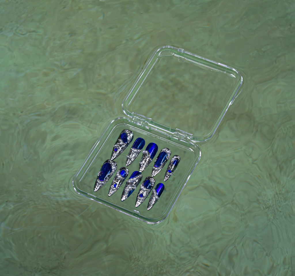 Set of blue and silver press-on nails in a clear plastic case on a greenish water-like background